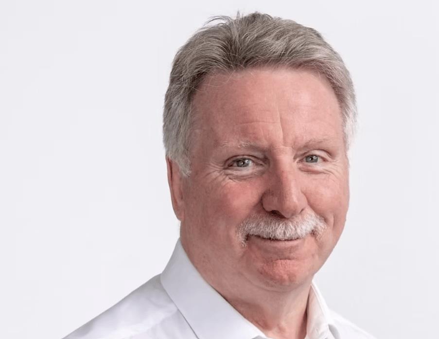 Wearing a white shirt, a grey-haired and moustached David Roberts smiles at the camera, in front of a white background.