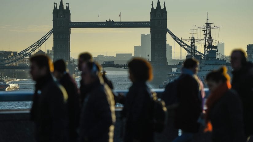 Silhouetted pedestrians walk on a bridge with Londons Tower Bridge and the River Thames in the background.