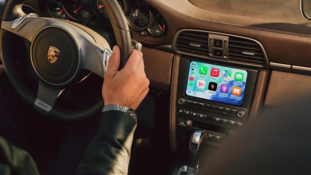Driver's hand on the steering wheel with a smartphone interface displayed on the car's dashboard screen.