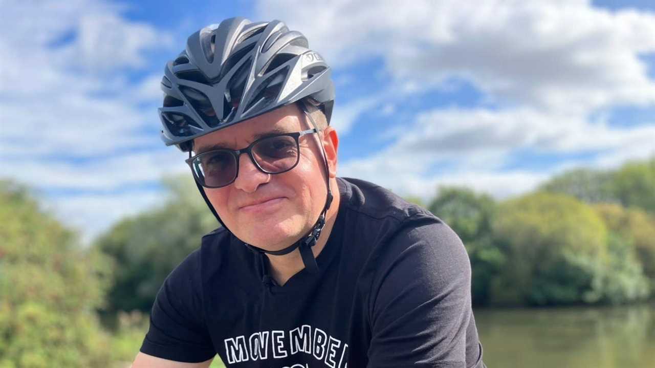 Ben Lidgey is wearing a grey cycling helmet and glasses. He is sat on a bicycle. Ben is wearing a black T-shirt with the