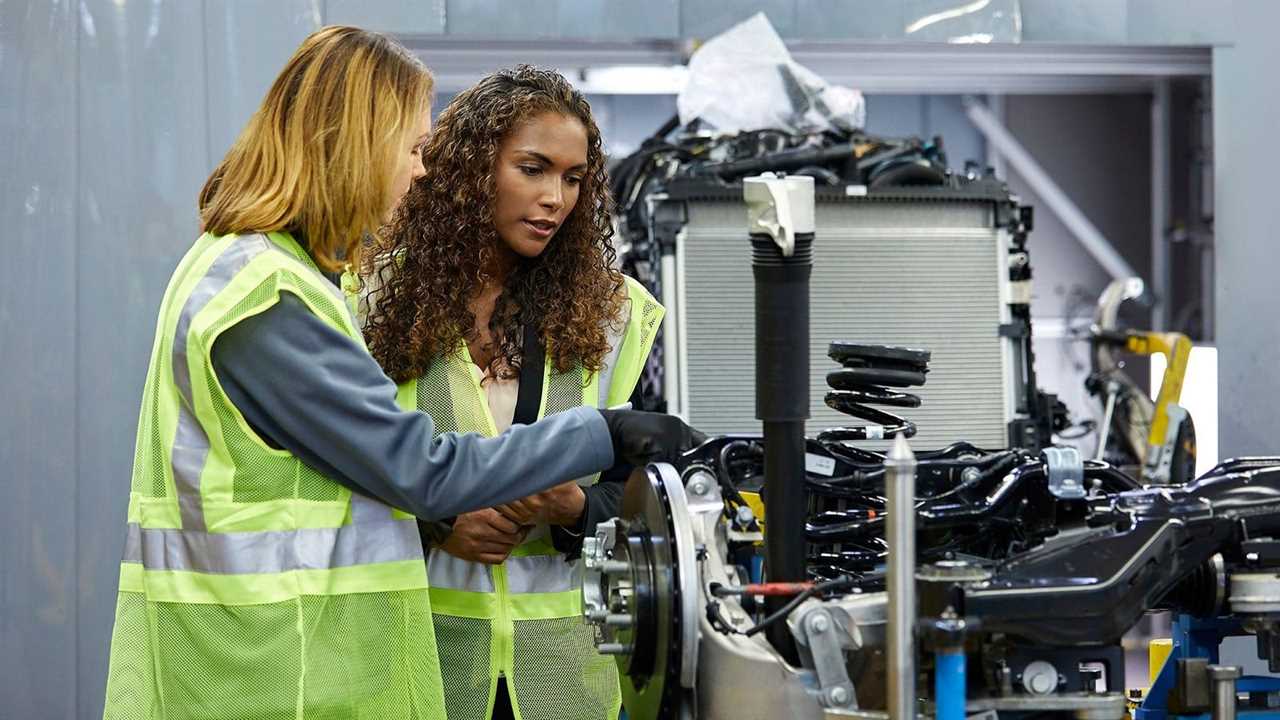 A stock image shows two people in reflective vests inspecting the chassis part of a car before assembly with the rest of